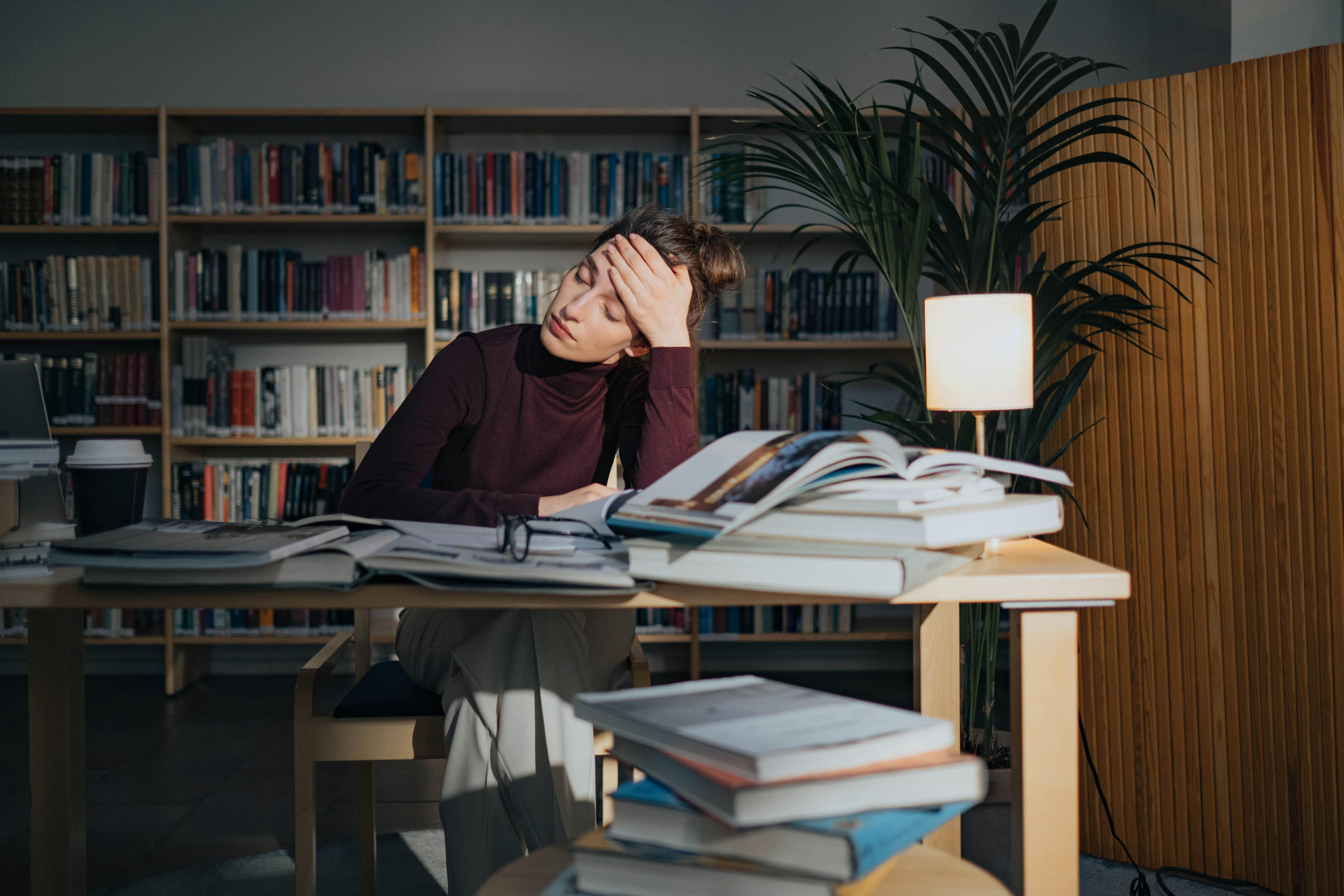 Tired student studying at a desk surrounded by open books and notes in a library.