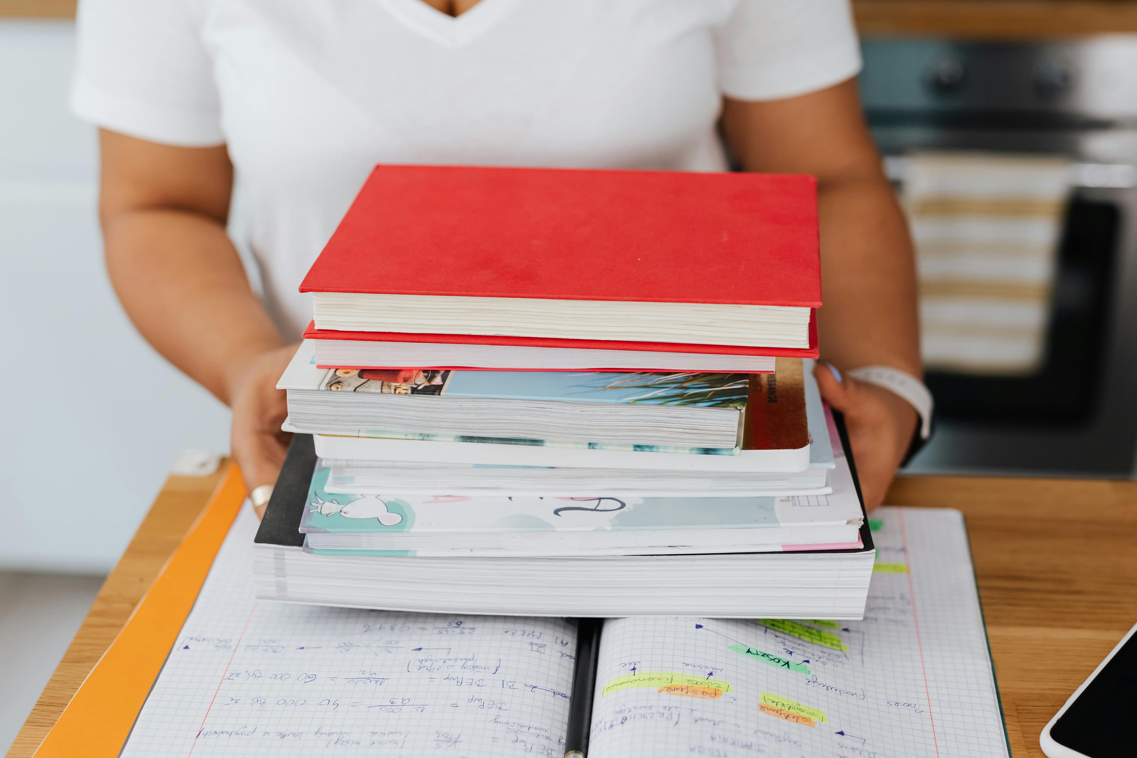 Person holding a stack of colorful textbooks over an open notebook filled with study notes.
