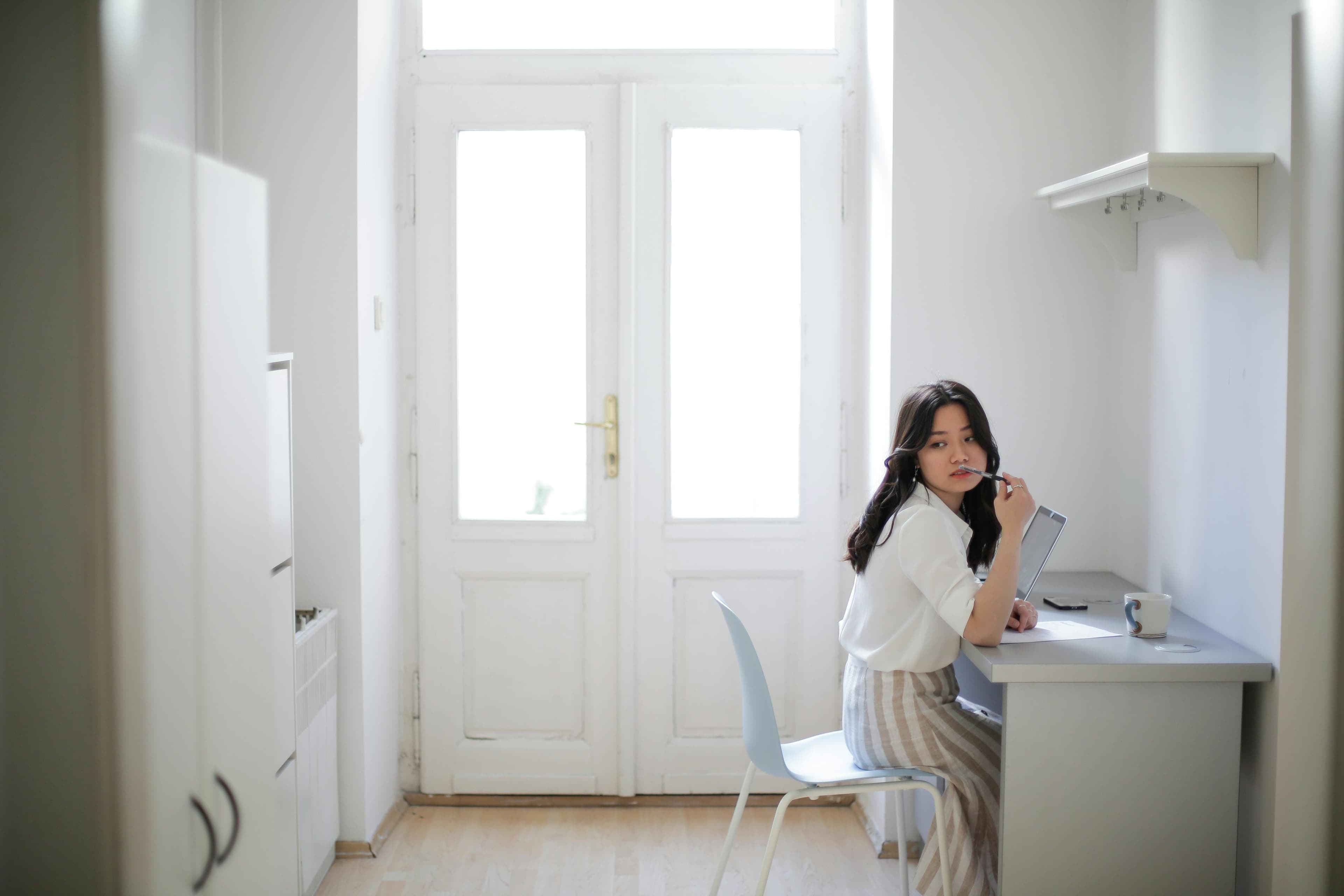 Student sitting at a desk in a bright room, thinking while holding a pen and studying with a laptop.